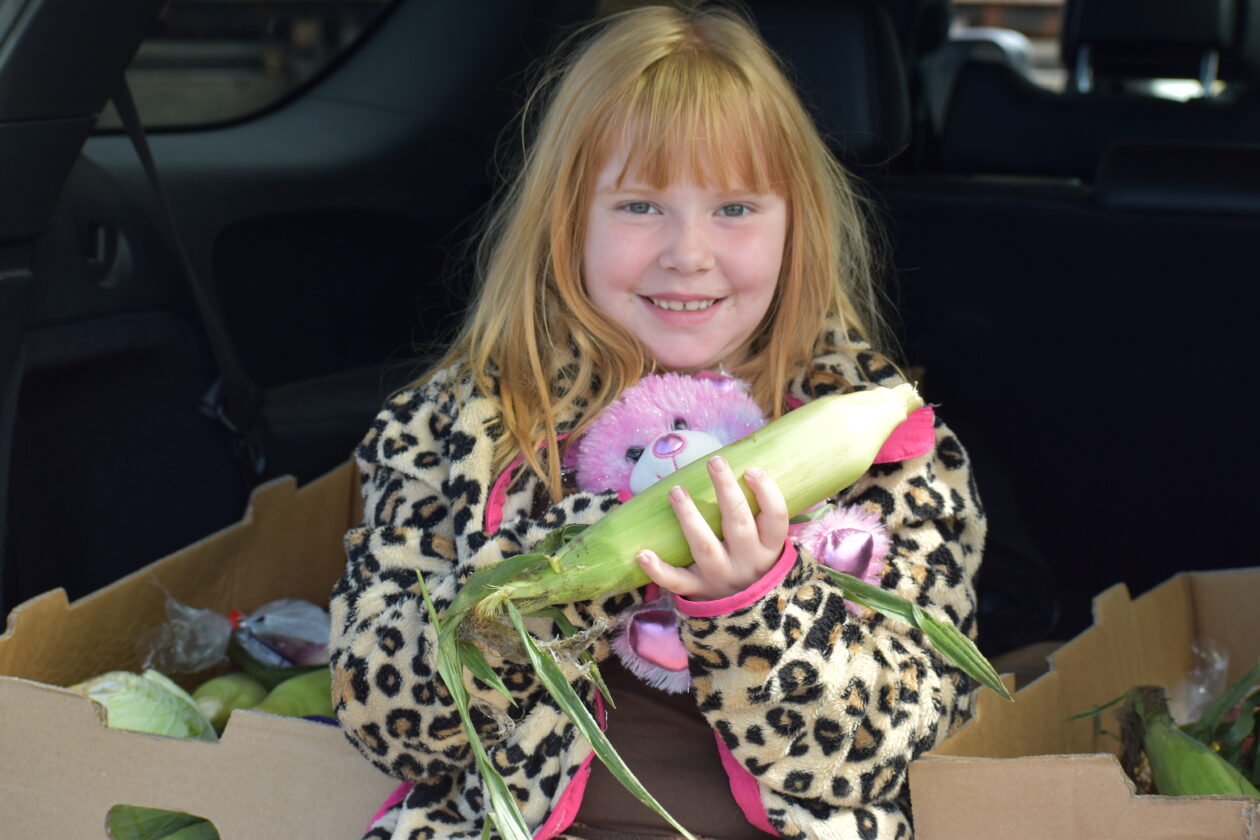 Katrina sits with her family's food in the back of her car holding a stuffed animal and ear of corn.
