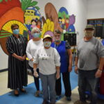 A few volunteers stand in front of a mural of fruit, bread and more at IM Kids' Warehouse