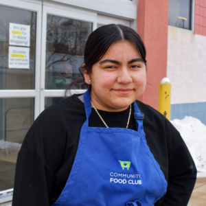 Maria poses in front of the food club wearing an apron