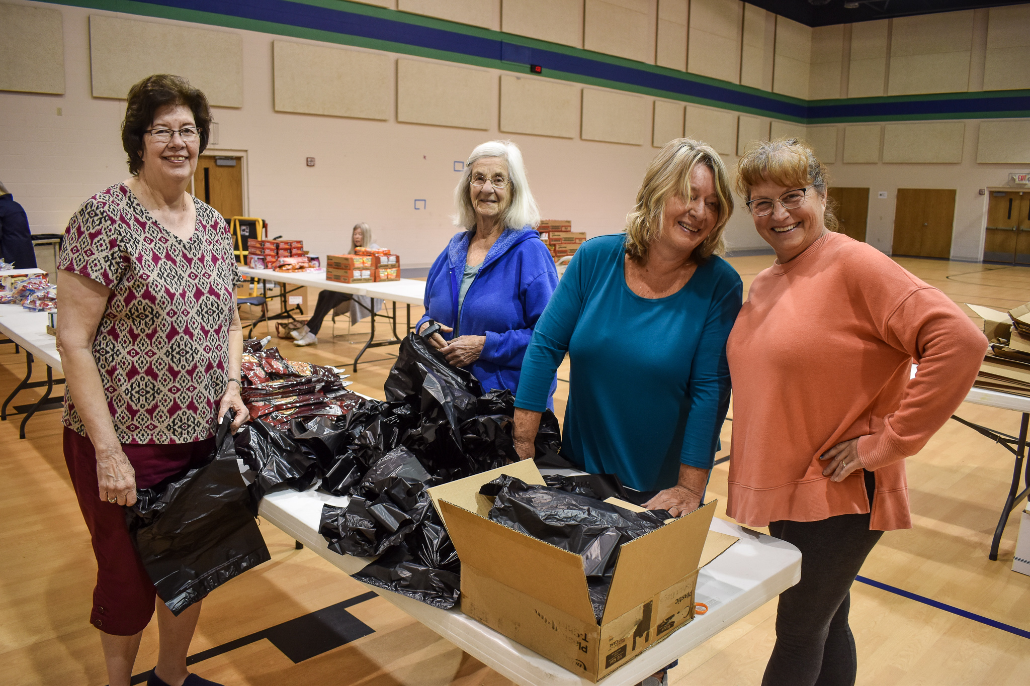 Four volunteers stand in a gym, posing as they fill bags with food