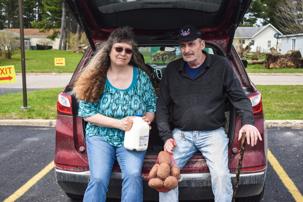 The couple sits on the back of their car holding milk and potatoes