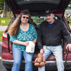 The couple sits on the back of their car holding milk and potatoes