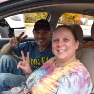 Michael (left) and Sarah (right) smile while sitting in their car and holding up peace signs with their fingers