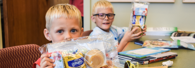 two boys sitting next to each other holding packaged meals while staring directly at the camera