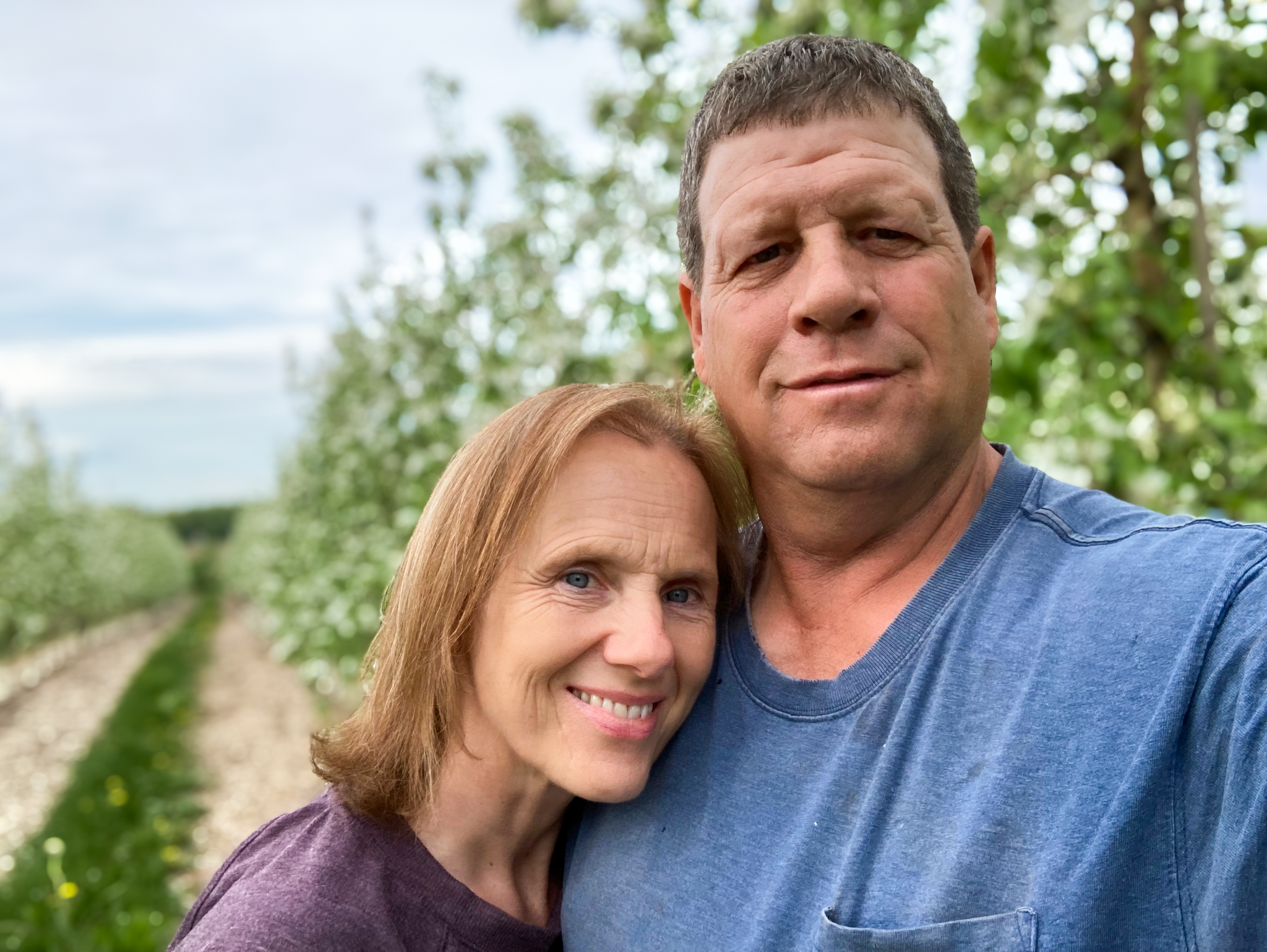 Deb and Duane Rasch smiling in front of an orchard.
