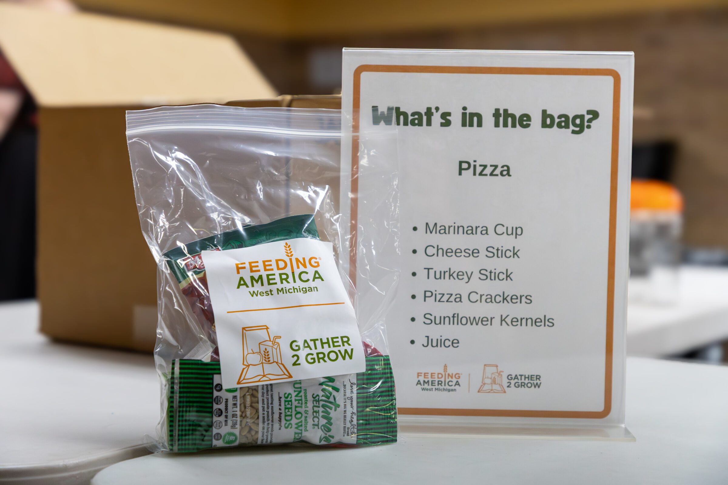 A Gather 2 Grow summer meal bag containing food items next to a display sign describing what's inside. 
