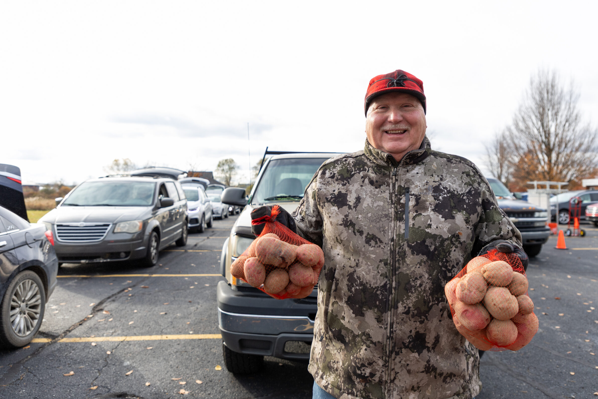 Mobile pantry volunteer at Church of the Savior in Coopersville smiling in front of a line of cars and holding bags of potatoes.