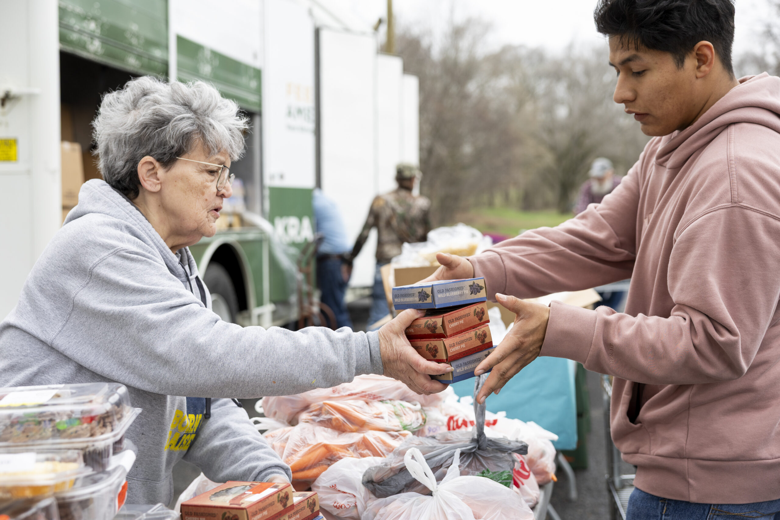 A woman passing small boxes of food items over to a young man while they stand by a table of food to be passed out.