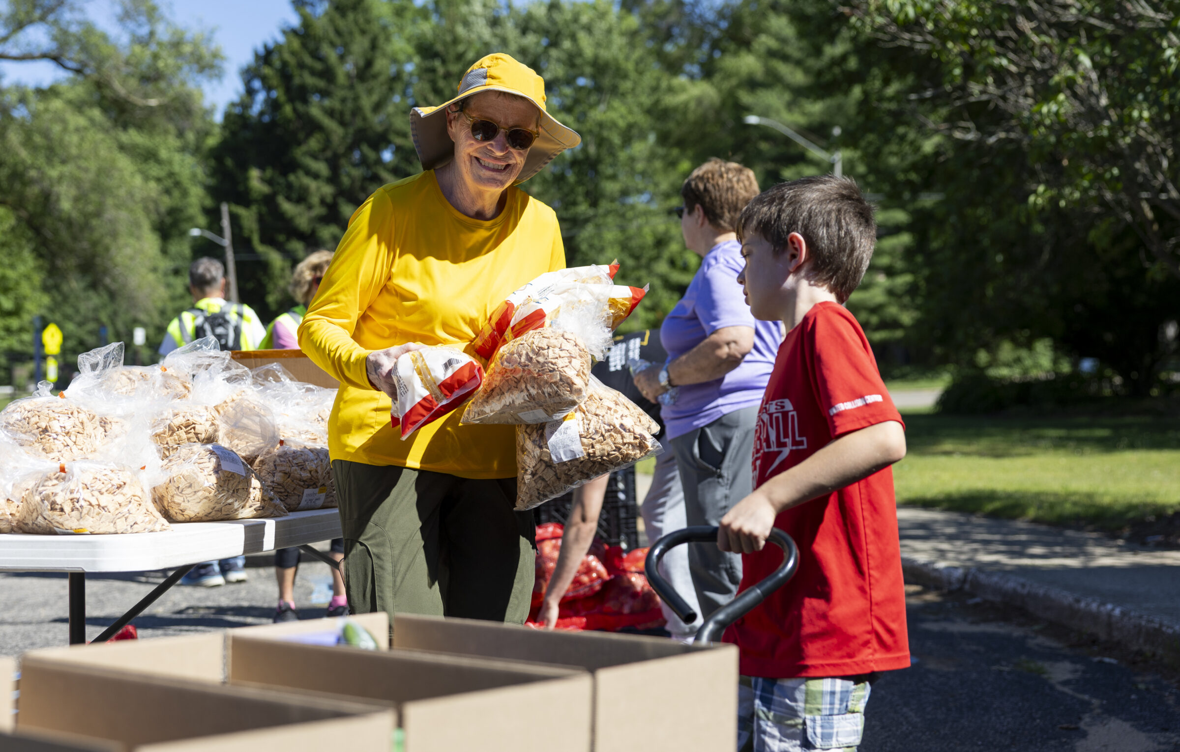 A woman holding bags of food and smiling. Next to her is a young boy  wheeling a cart with several boxes. 