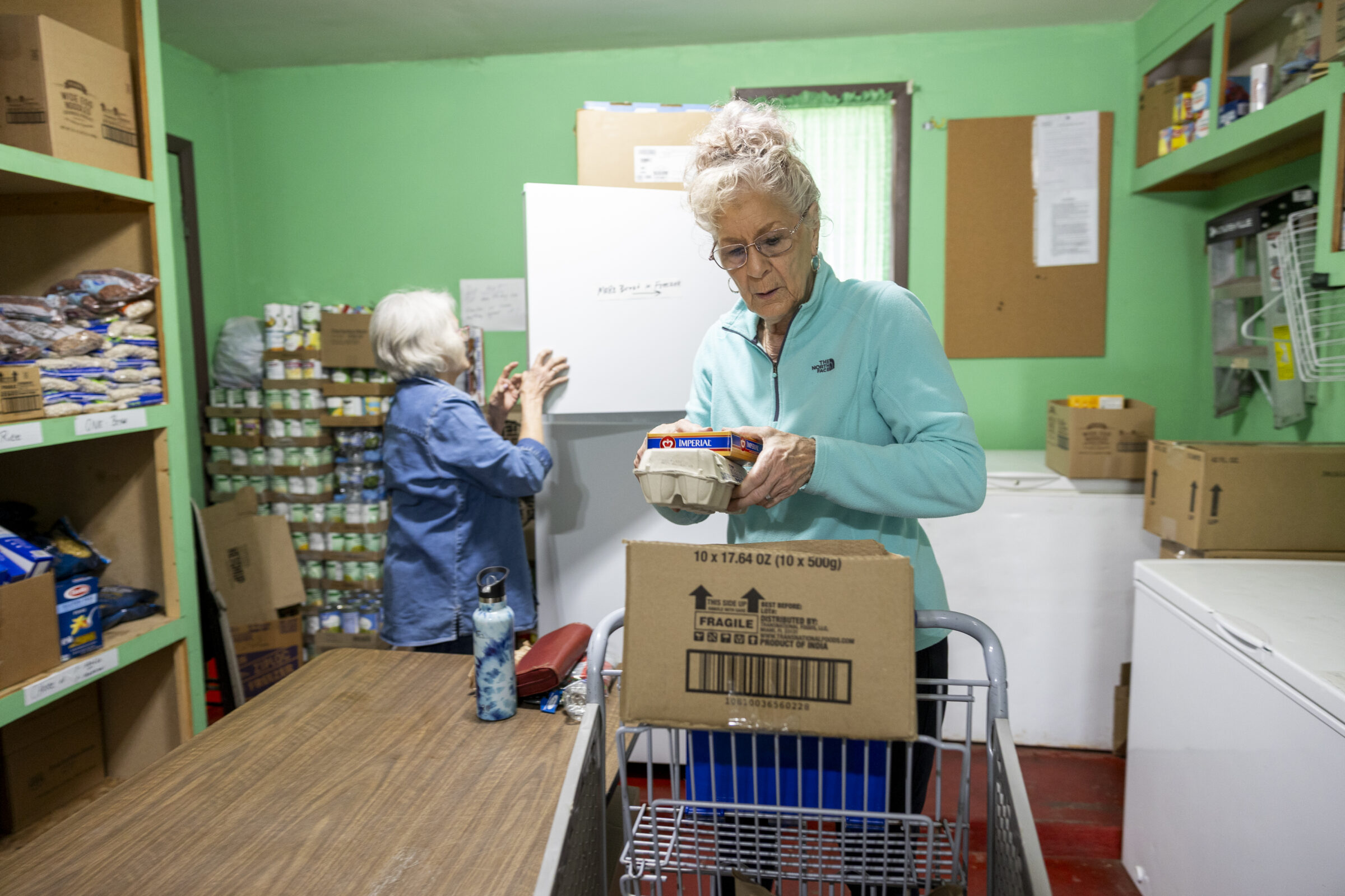 Two women who volunteer at the pantry. One woman fills a box with food items and the other looks through a pantry freezer. 