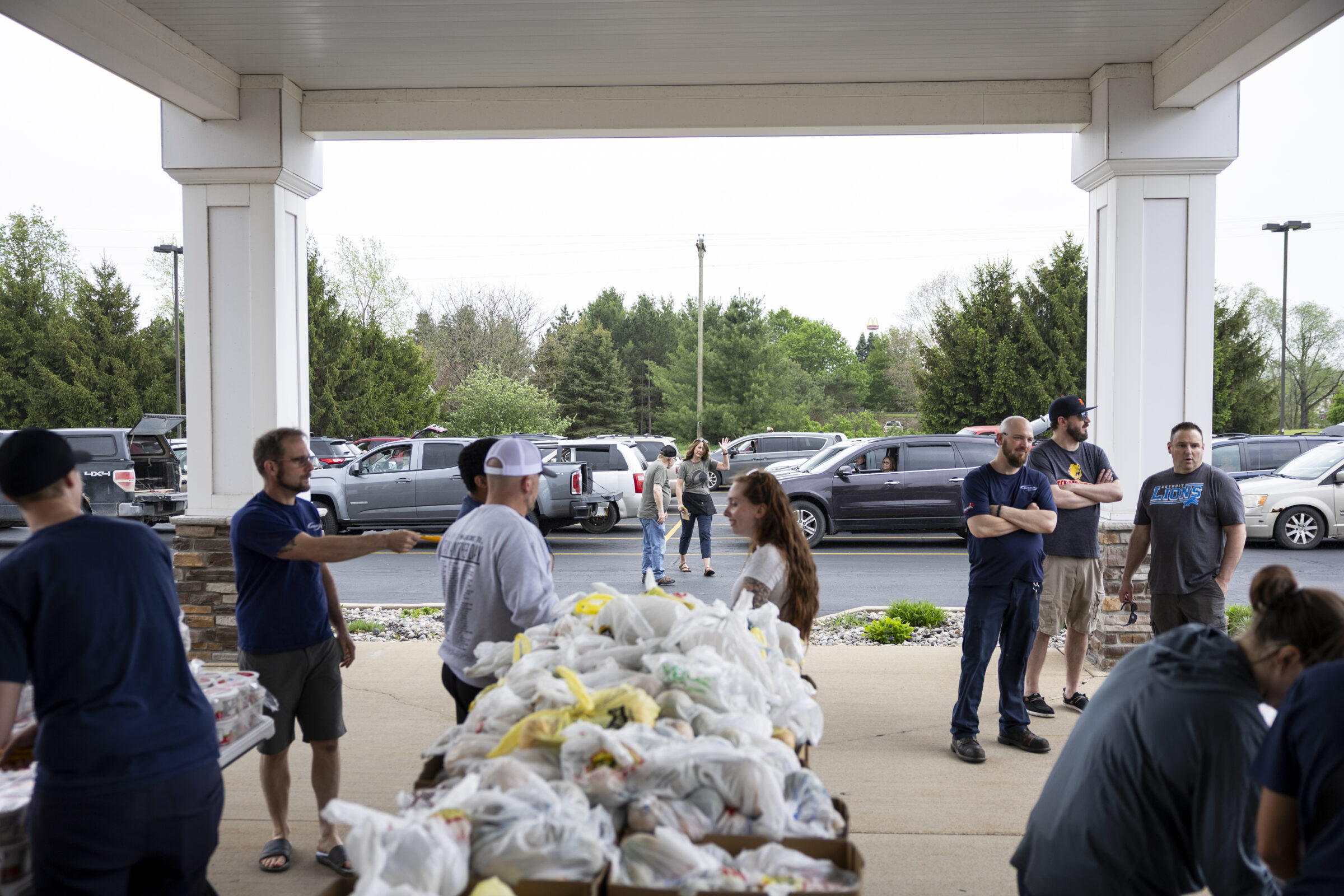 Volunteers by a table stacked with boxes of grocery bags, whcih contain various food items. In the background, there is a line of cars.