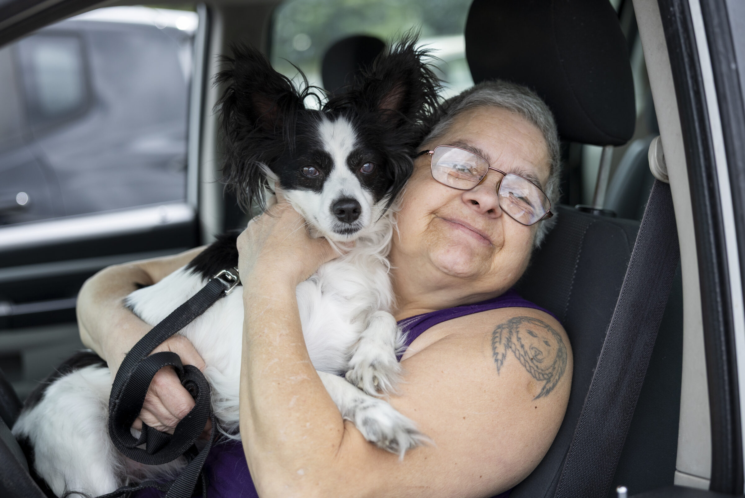 A woman in her car smiles while holding her small black and white dog close.