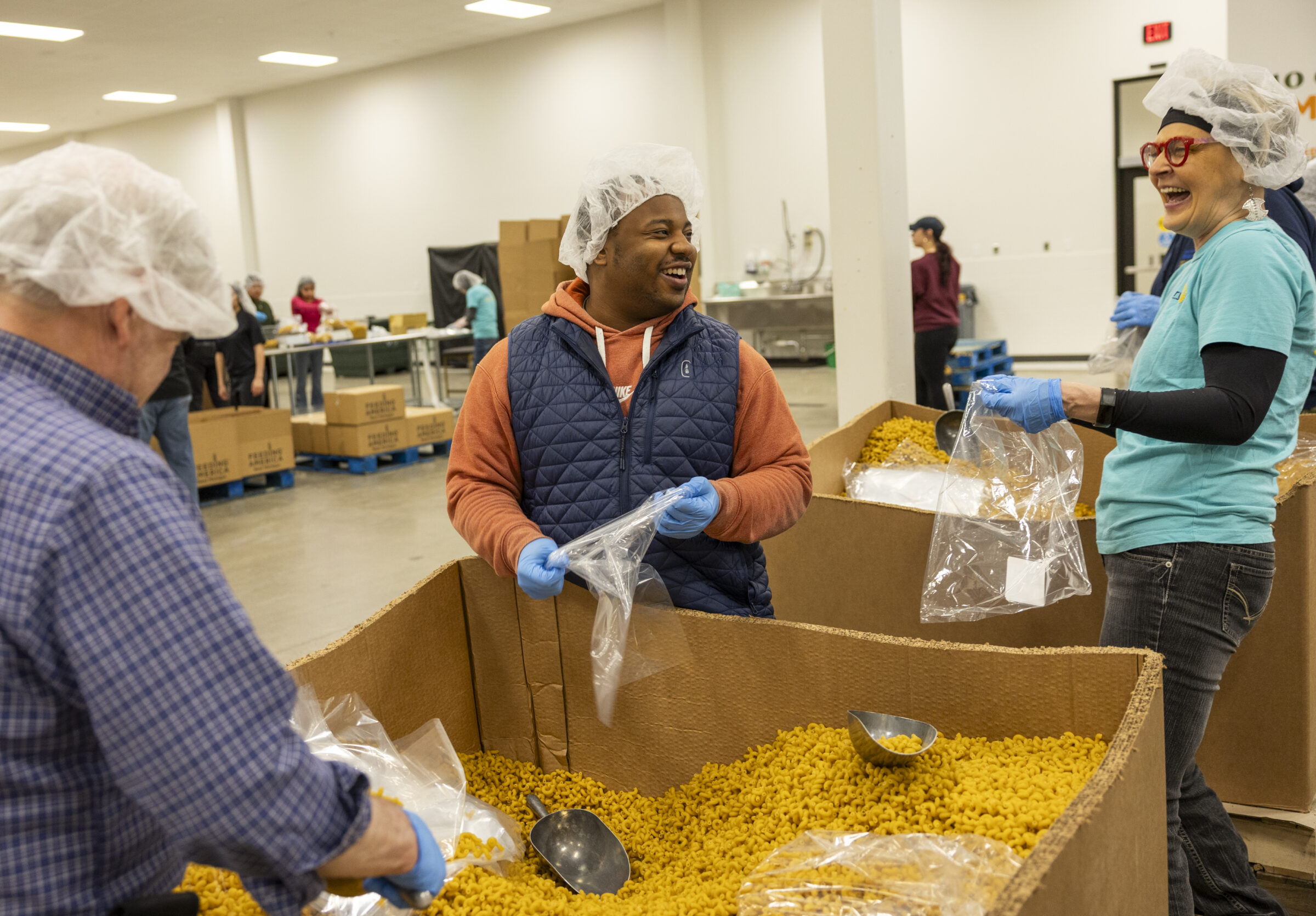 Two volunteers laughing while standing by a large cardboard tote filled with pasta noodles. 