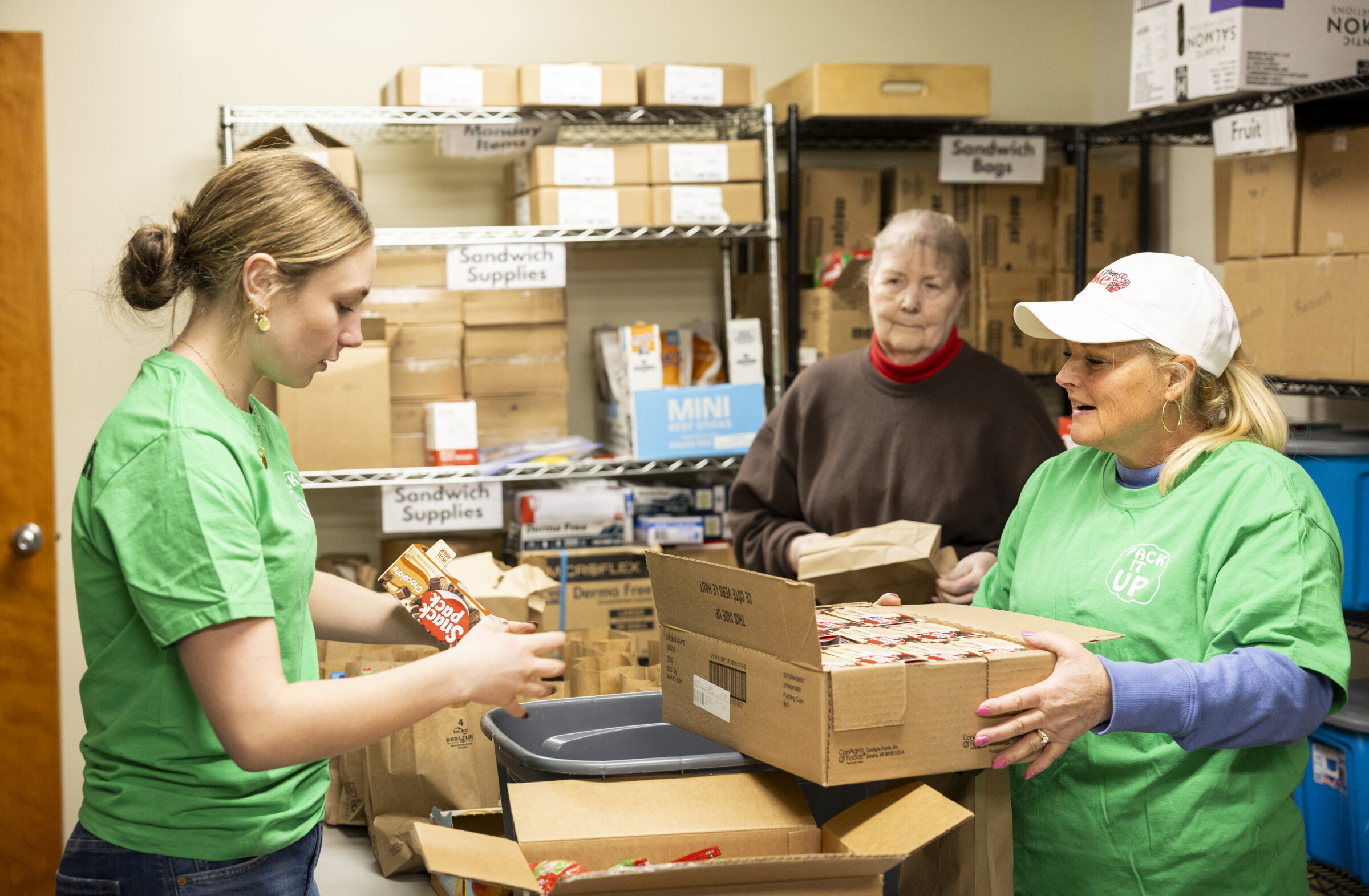 Three volunteers stand around boxes of lunch snacks, sorting through them and putting them in paper lunch bags. 