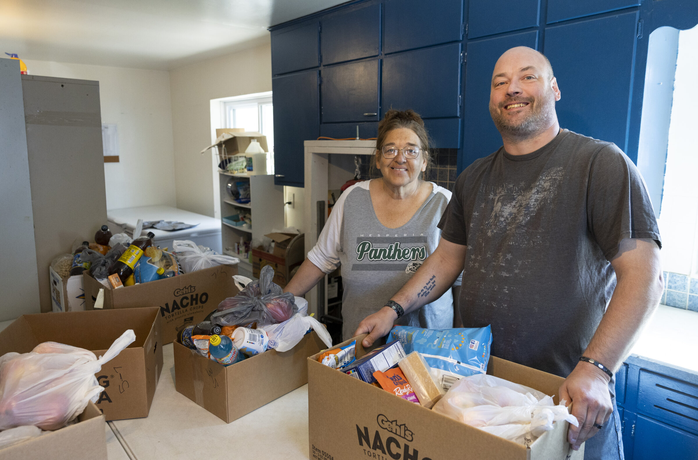 A man and a woman smiling while standing in front of a table with boxes. In the boxes are various packaged food items.