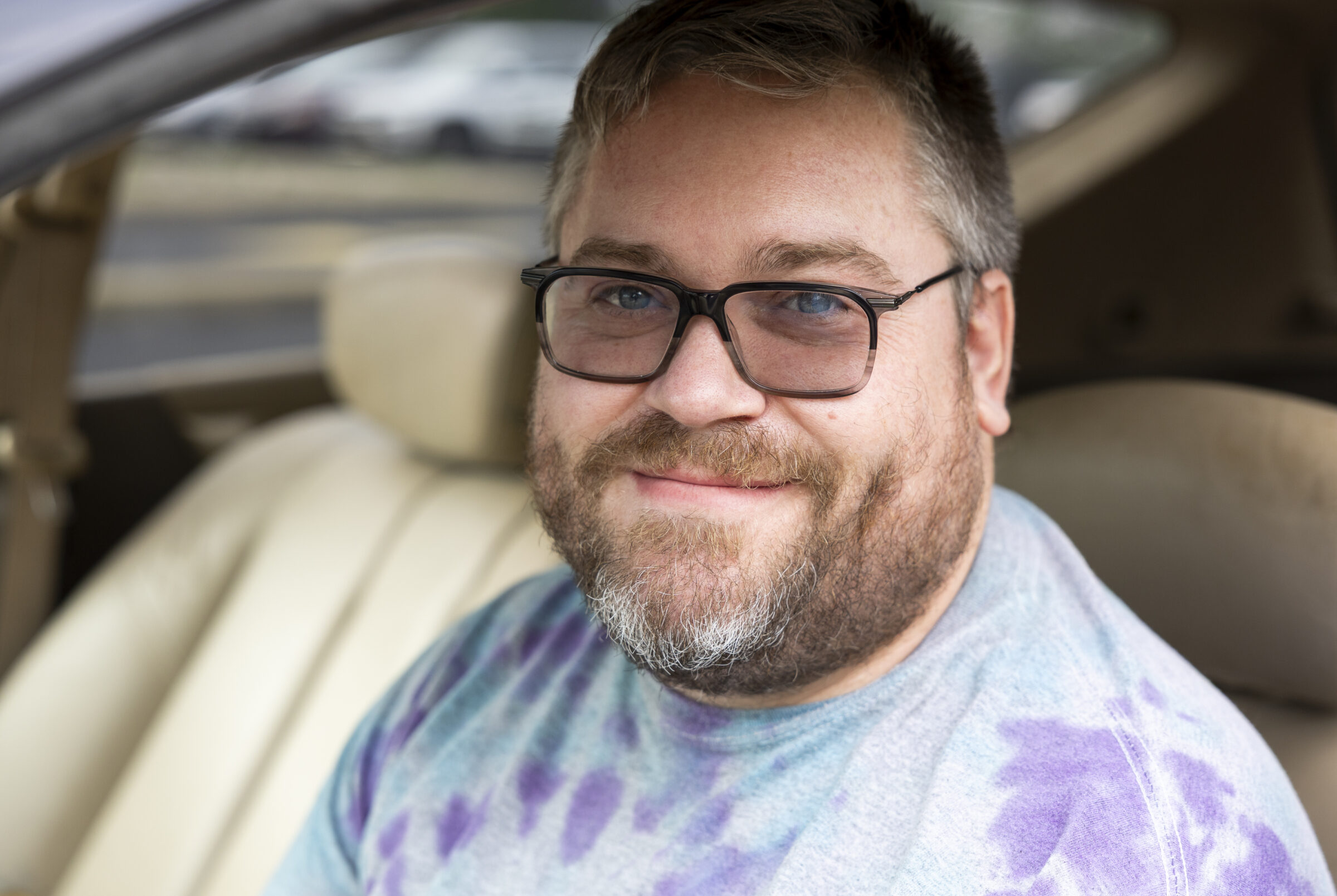 A man wearing glasses and a tie dye shirt, sitting in his car and smiling.