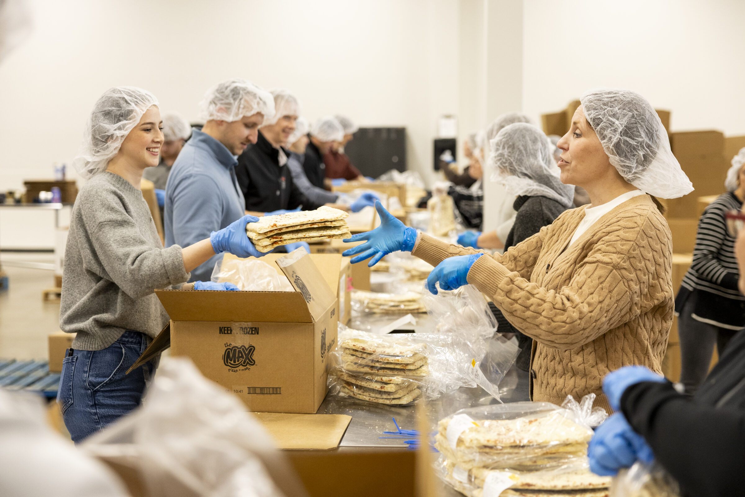 Several Meijer volunteers standing around long metal tables. There are boxes and frozen pizza in clear bags on the table. One volunteer is passing to another a stack of small frozen pizzas.