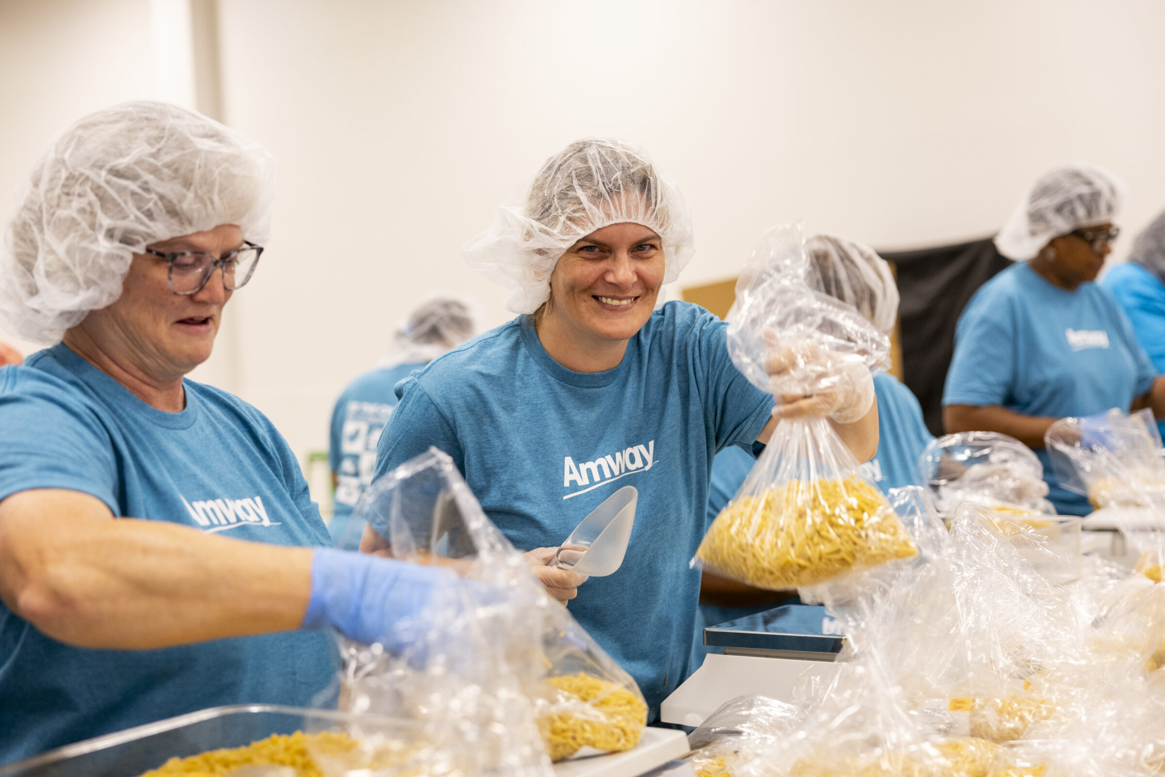 Several volunteers in Amway shirts smiling and standing at a table. The table is filled with scales and bags of noodles.