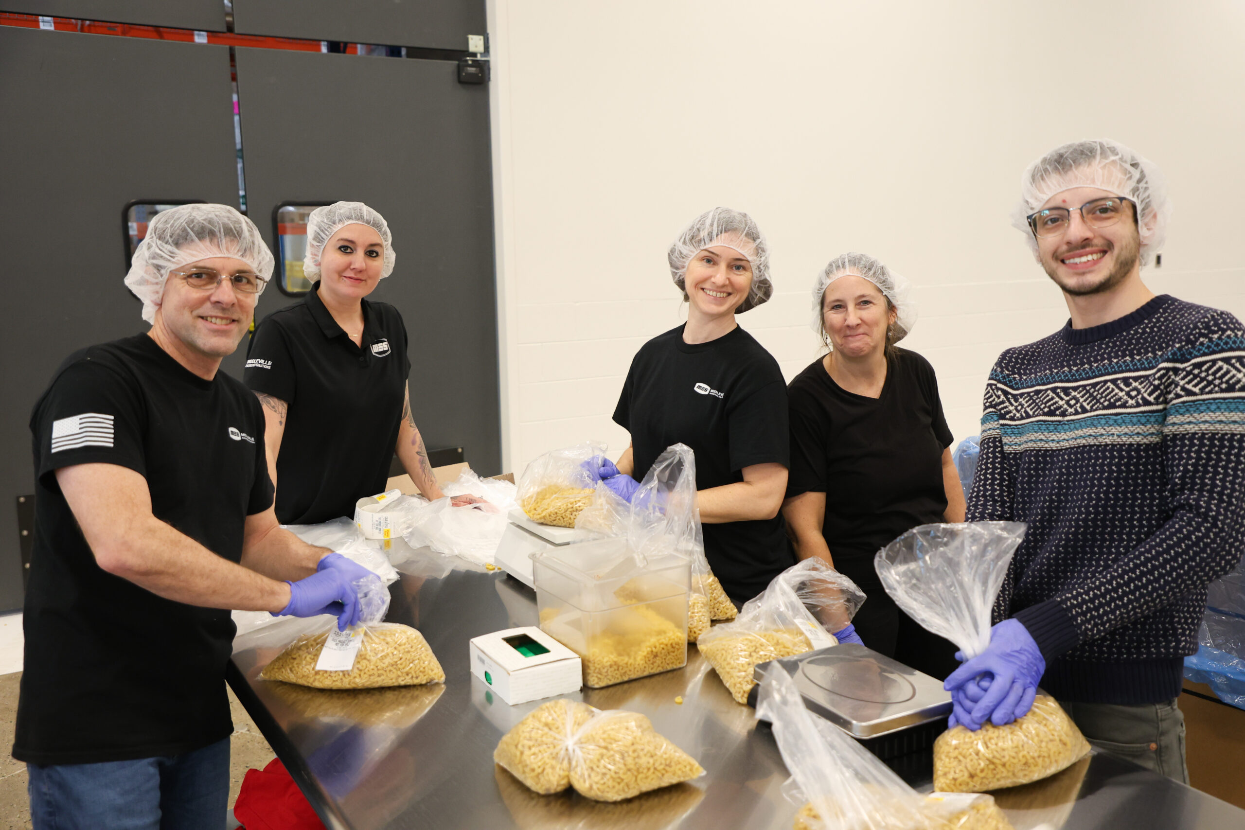 Five people standing around a metal table, smiling. There are scales, clear bins, and bags of pasta on the table.
