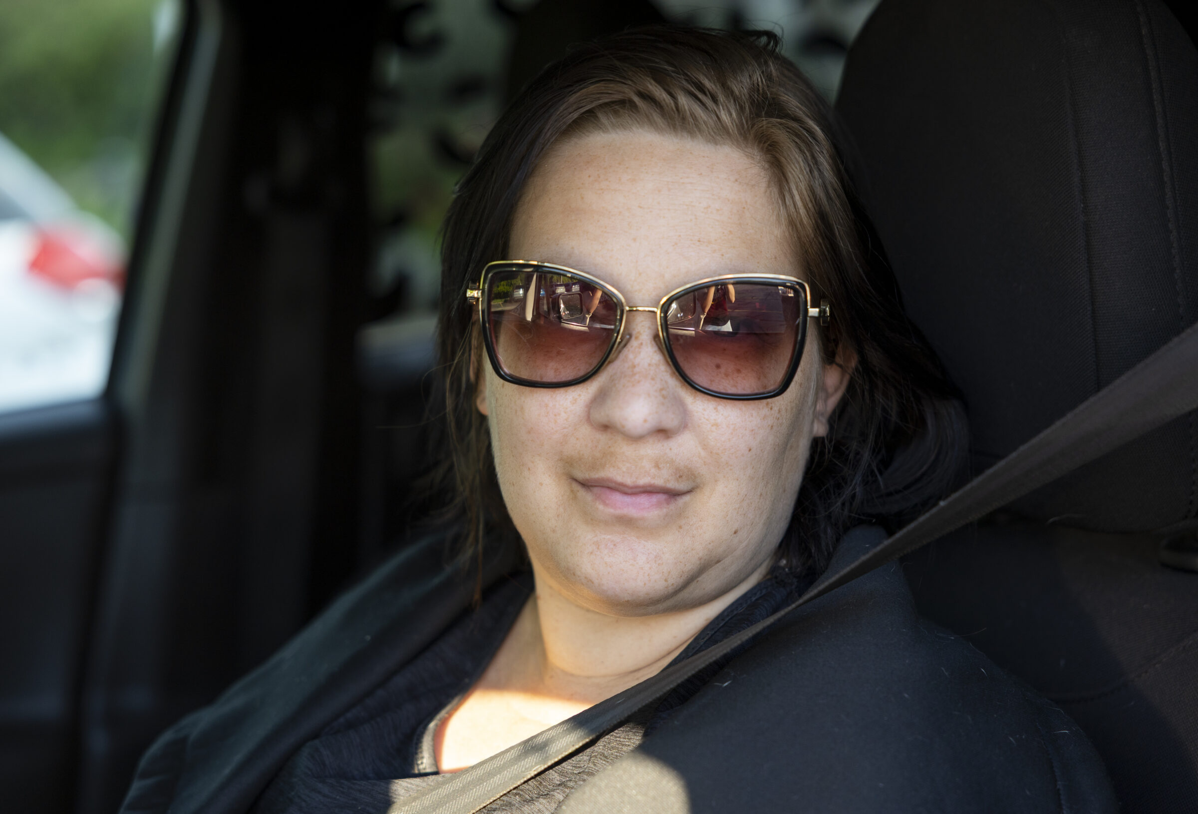 A woman wearing sunglasses sits in her car while looking at the camera.
